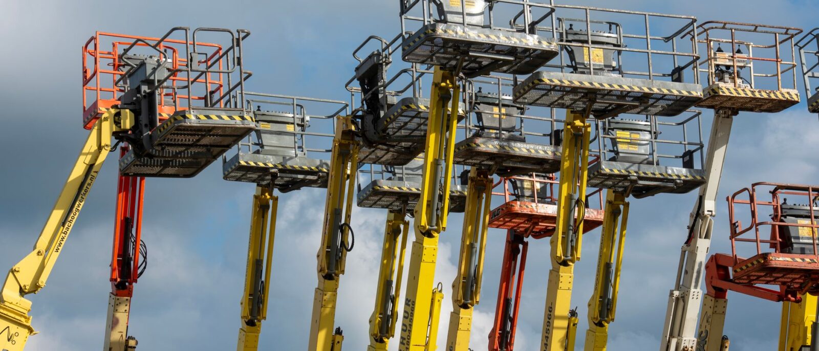 Multiple aerial work platforms with colorful booms reaching skyward against a cloud-dotted backdrop.