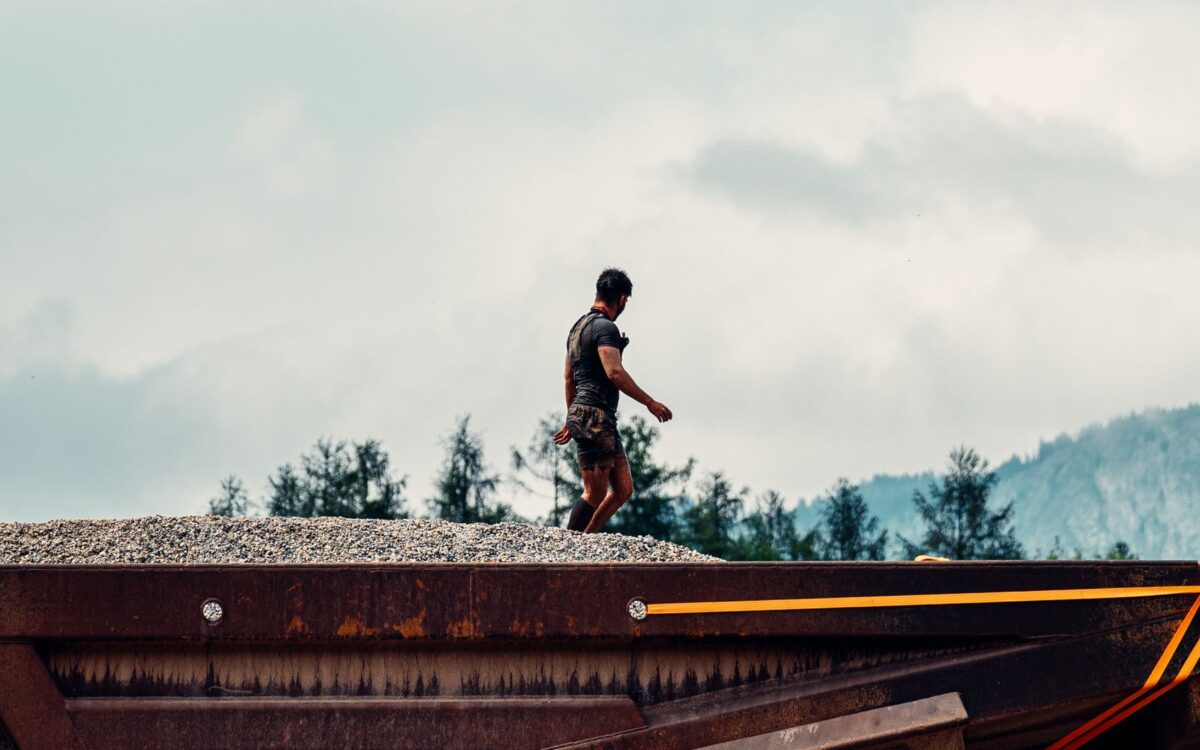 A construction worker walking on a gravel heap with mountains and trees in the background.