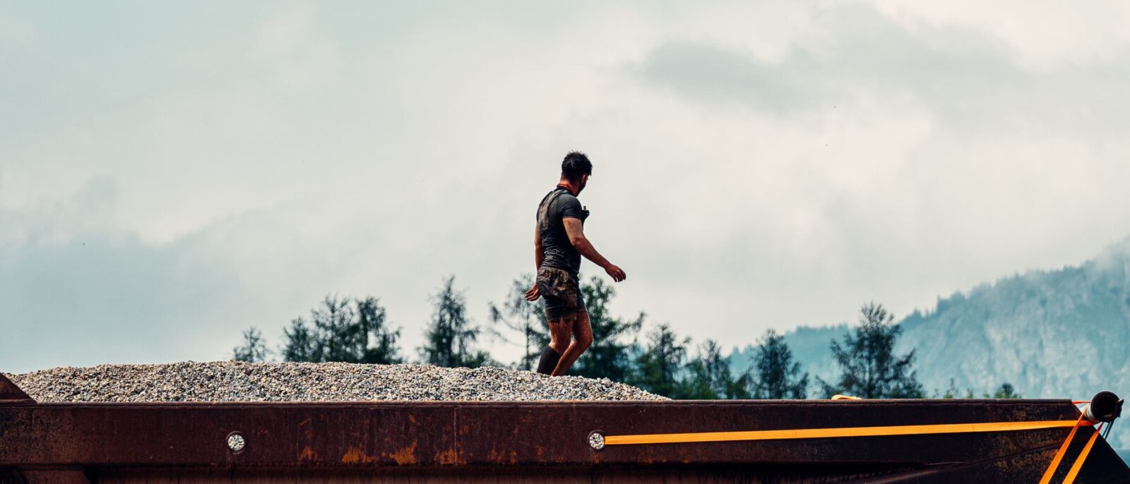 A construction worker walking on a gravel heap with mountains and trees in the background.