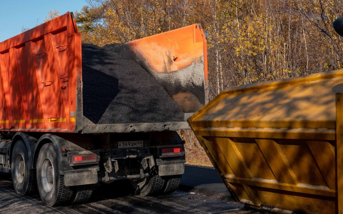 A dump truck unloading asphalt into a construction bin on a sunny autumn day.