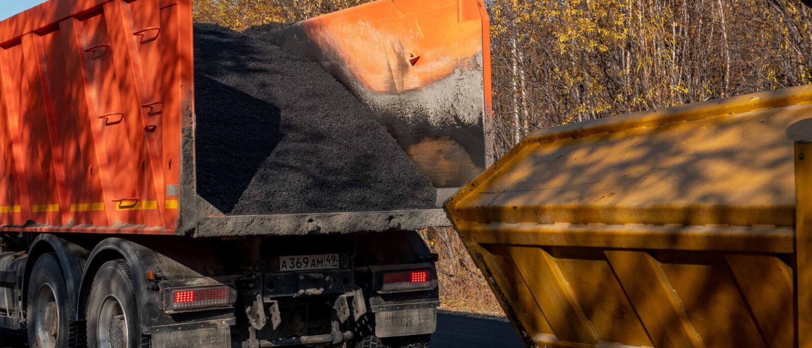 A dump truck unloading asphalt into a construction bin on a sunny autumn day.