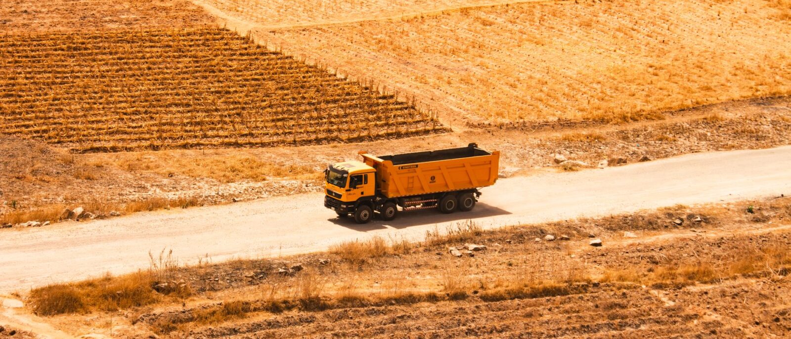 Aerial view of an orange dump truck on a rural road surrounded by dry fields and agricultural land.