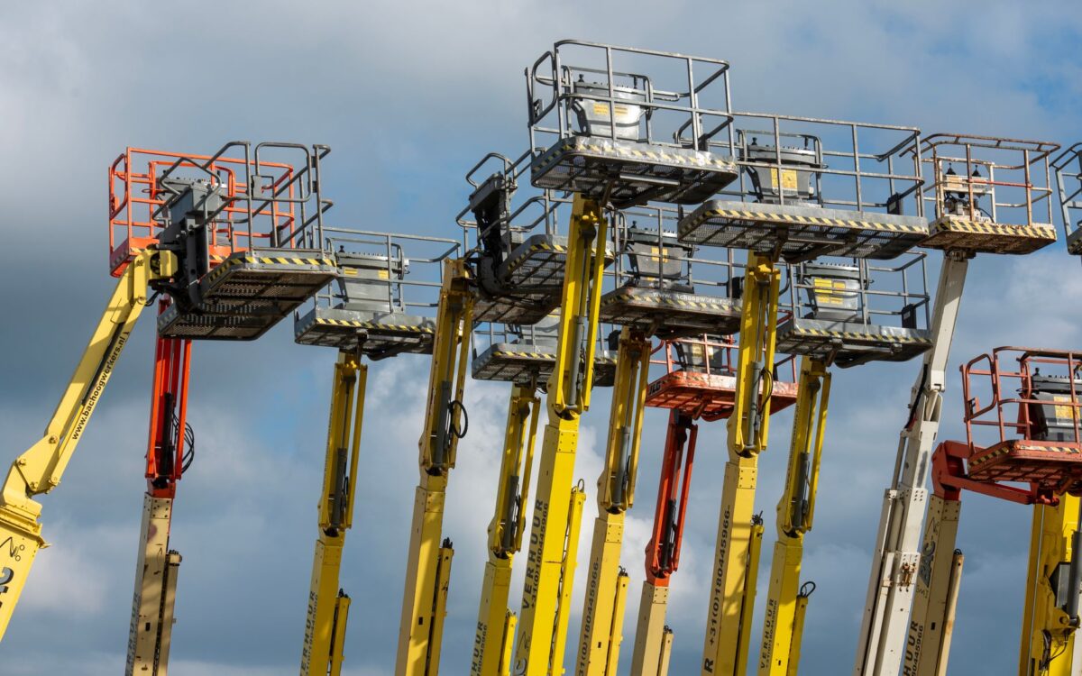 Multiple aerial work platforms with colorful booms reaching skyward against a cloud-dotted backdrop.