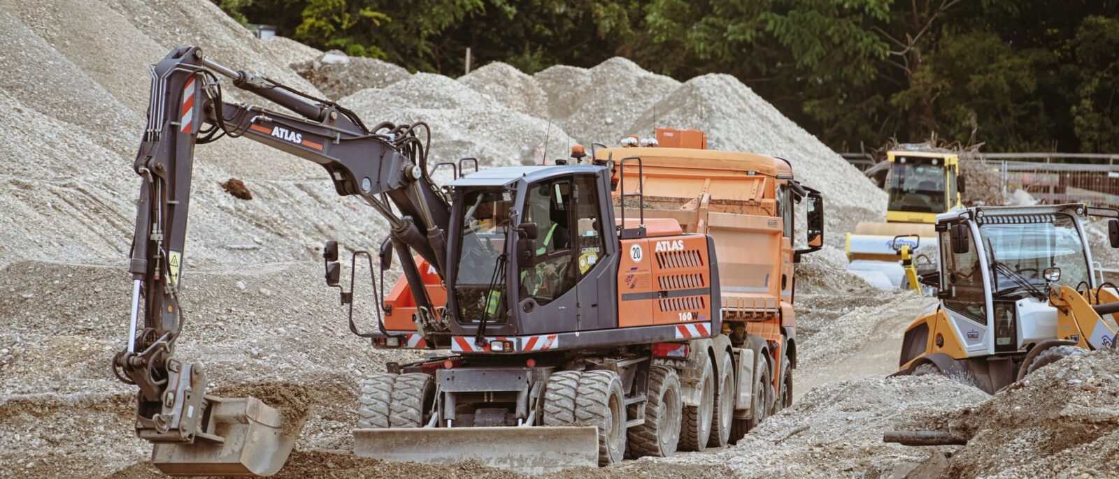 Excavator and machinery at a bustling construction site with piles of sand.