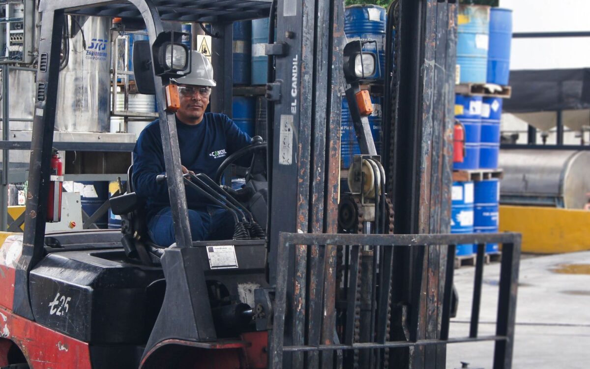 Warehouse scene with worker operating a forklift among blue barrels.