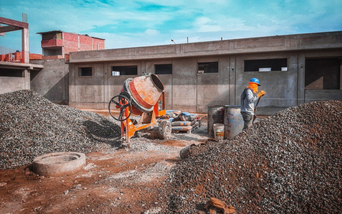 A construction site with a worker wearing safety gear next to a cement mixer and gravel piles.
