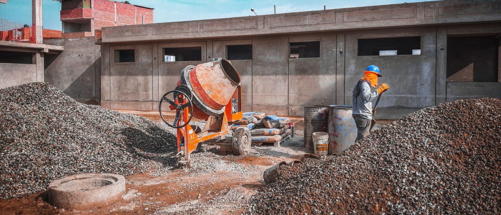 A construction site with a worker wearing safety gear next to a cement mixer and gravel piles.
