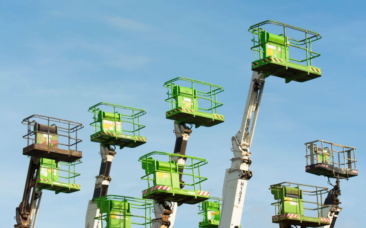 A group of green cherry pickers extending against a clear blue sky in an industrial setting.