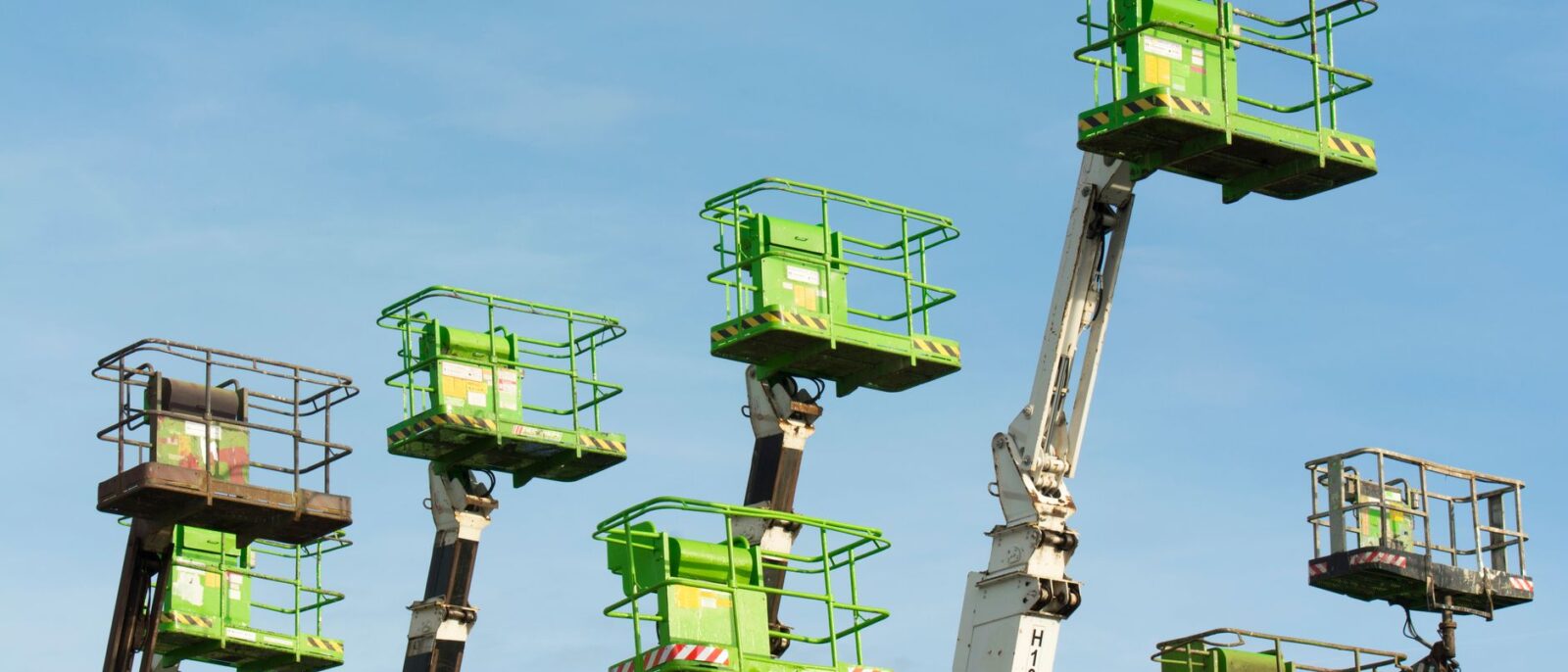 A group of green cherry pickers extending against a clear blue sky in an industrial setting.