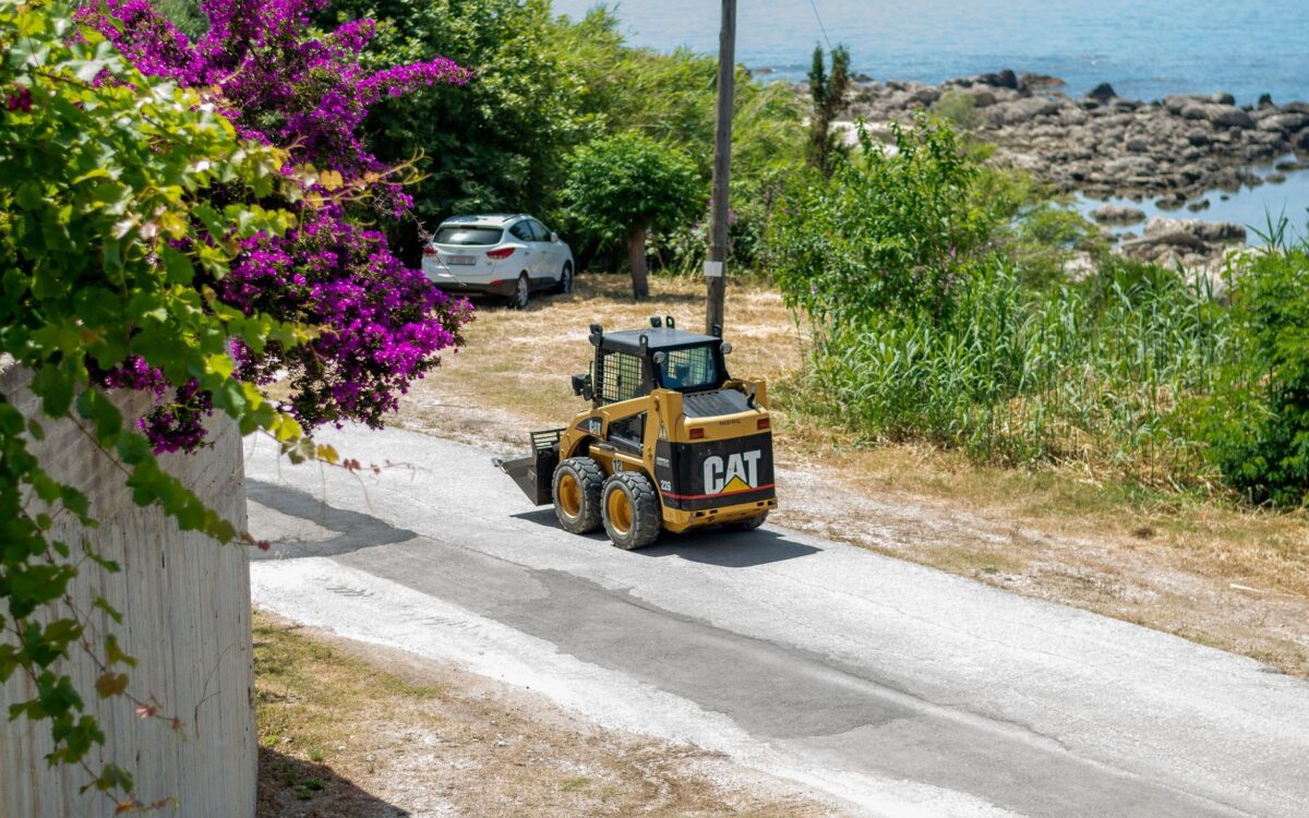 Skid steer loader on a sunny coastal road with vibrant foliage and sea view.