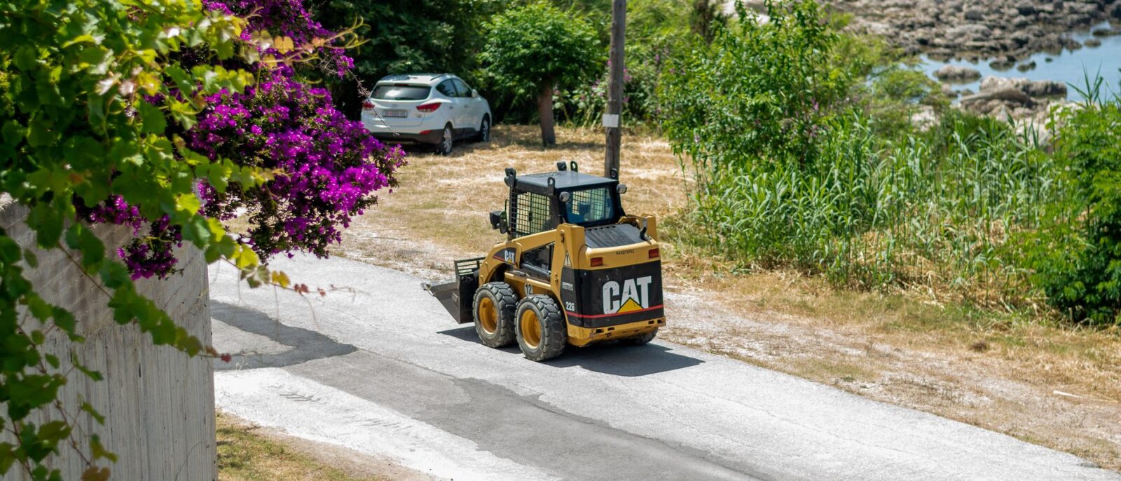 Skid steer loader on a sunny coastal road with vibrant foliage and sea view.