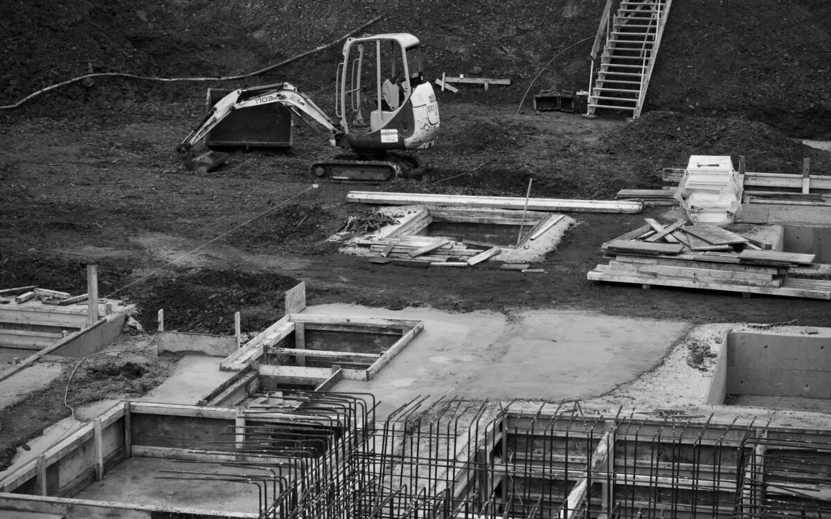 Monochrome image of an excavator at a construction site with foundations laid and rebar in place.