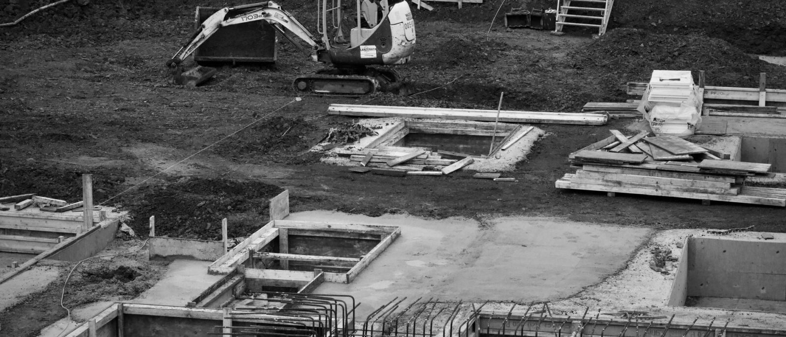 Monochrome image of an excavator at a construction site with foundations laid and rebar in place.
